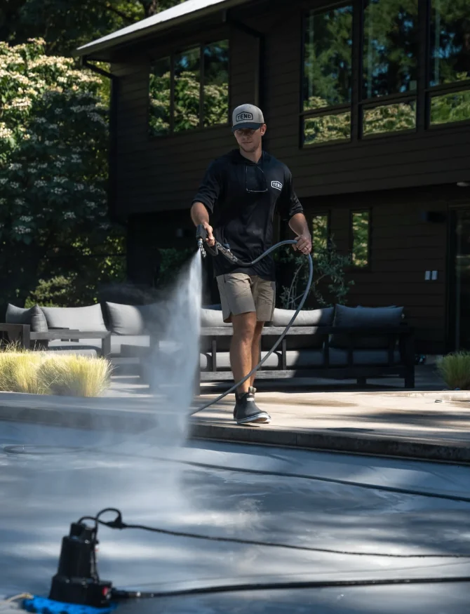 Tend technician pressure washing a pool deck in front of a dark PNW home surrounded by trees